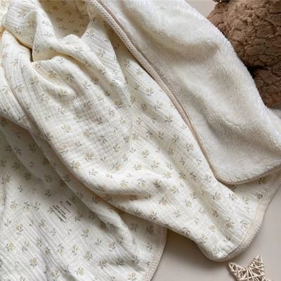 Couverture bébé blanche à motif floral champêtre drapée naturellement sur surface claire, mettant en valeur la douceur du tissu double face et les finitions délicates