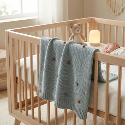 Couverture bébé bleu-gris tricotée suspendue sur lit à barreaux en bois naturel, bébé endormi paisiblement avec doudou ourson.