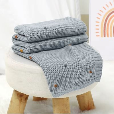 Couverture bébé bleu clair tricotée empilée sur tabouret scandinave blanc et bois, texture maille ajourée visible
